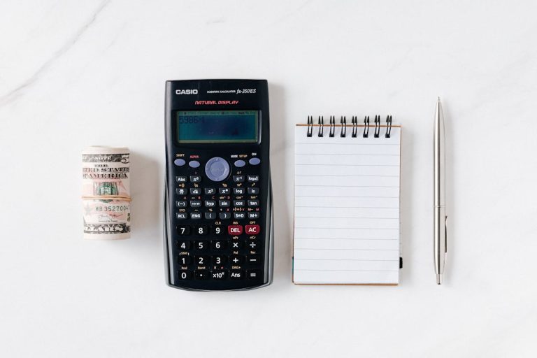 A roll of dollar bills next to a calculator and a notepad with pen